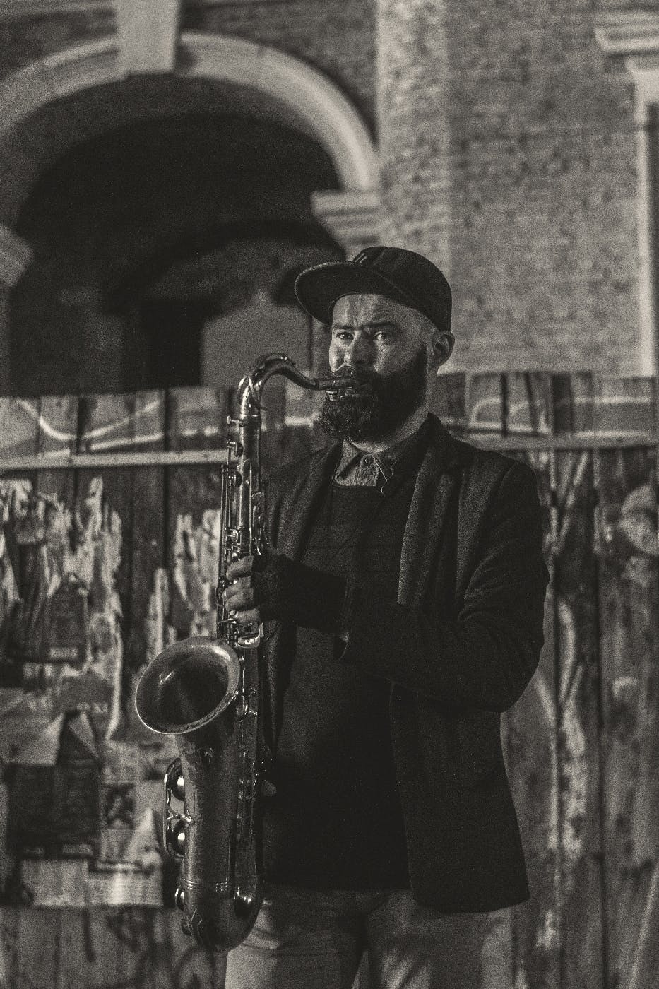 A bearded black man in ball cap, jacket and fingerless gloves playing sax in front of a graffitied wood fence