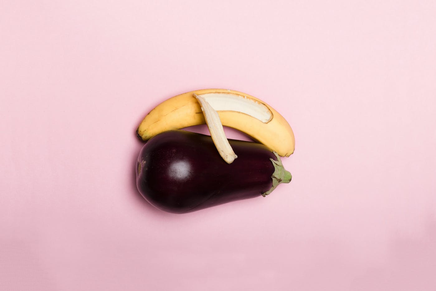 A banana and an eggplant side by side, a part of the banana's peel is draped on the ggplant like and arm