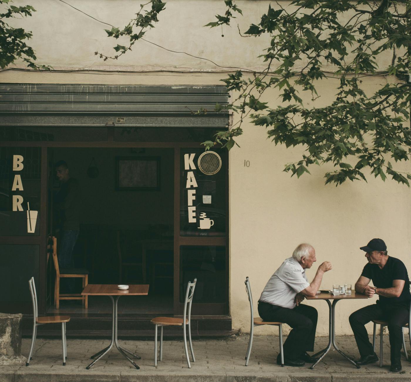 two men sitting at an outdoor table to the right of a bar/kafe