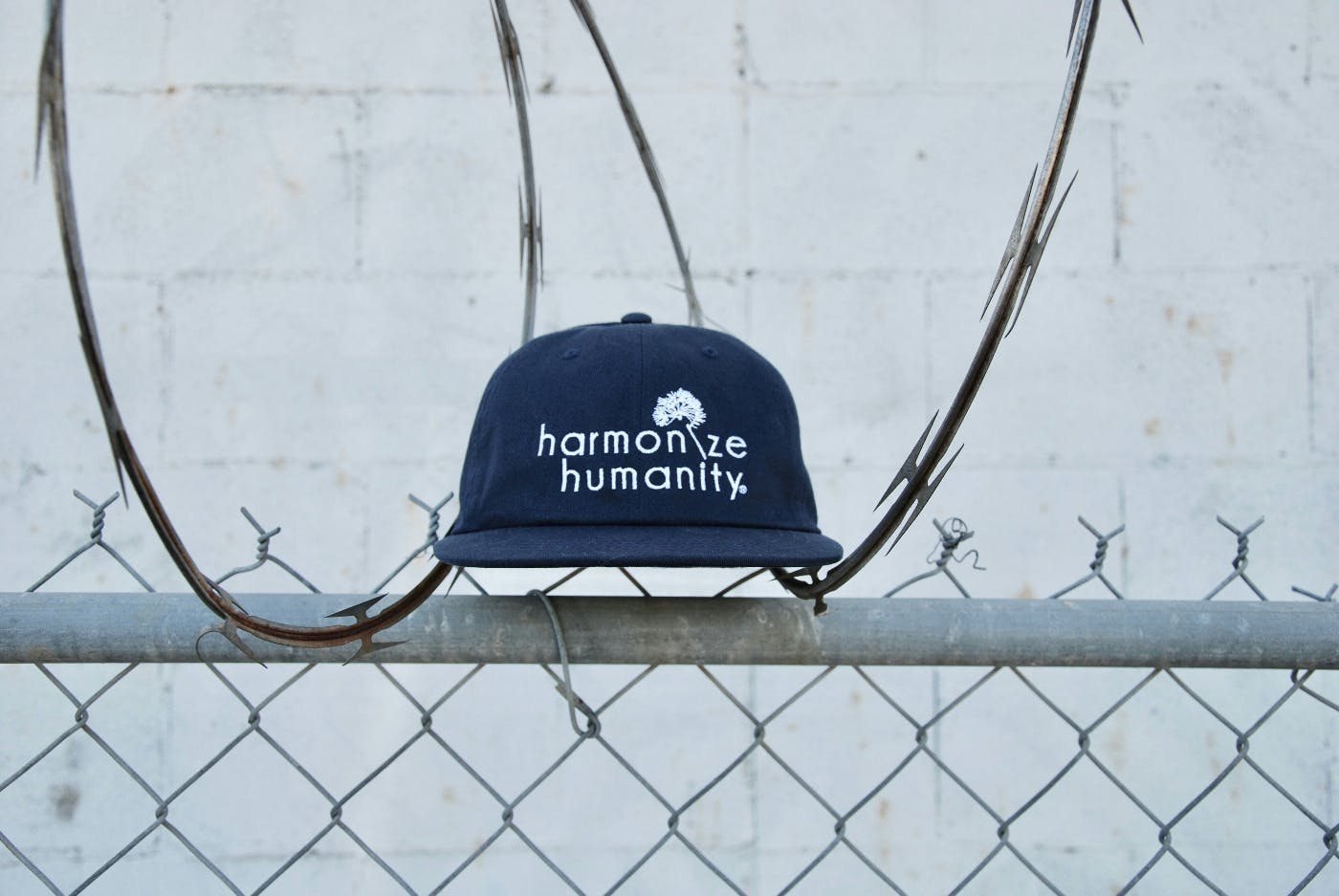 A blue baseball cap with Harmonize Humanity on it sitting on top of a fence covered with razor wire