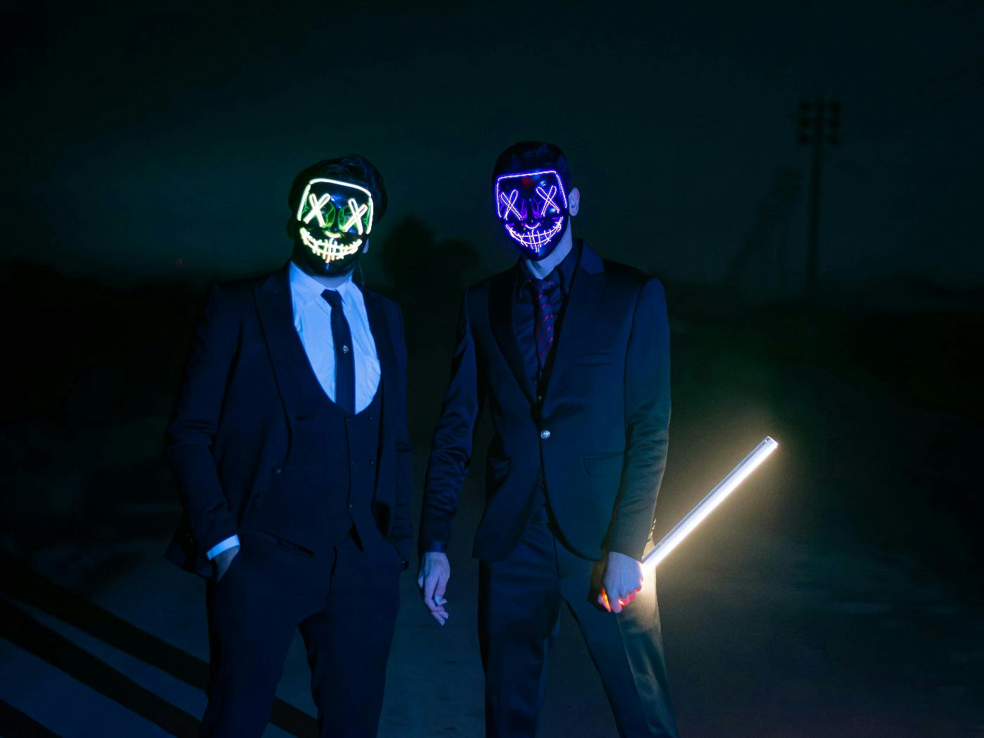 Two guys in dark suits wearing neon light masks, one holding a light stick