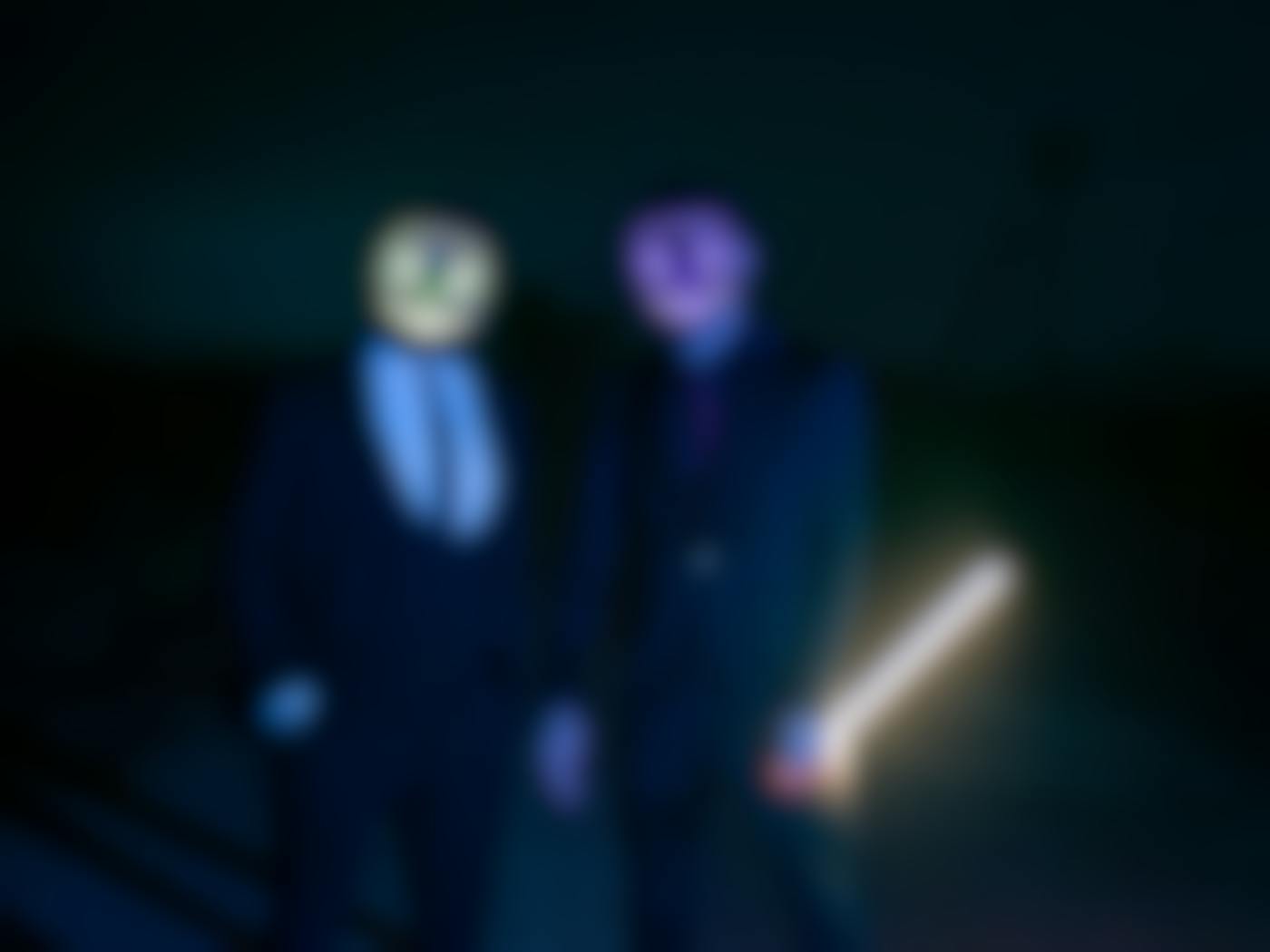 Two guys in dark suits wearing neon light masks, one holding a light stick