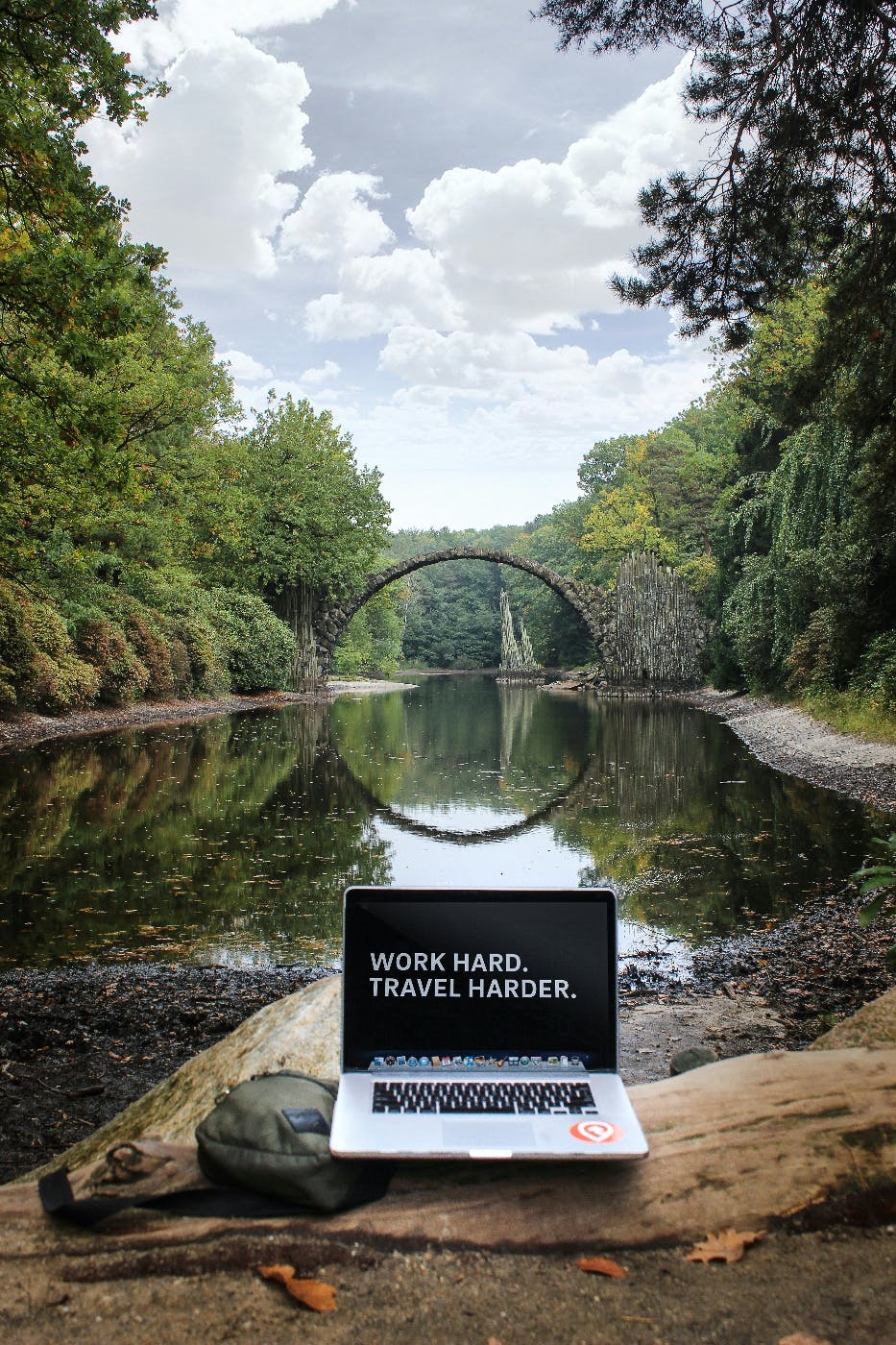 A river with an arched bridge reflecting in the water creating a circle, and a laptop in the foreground with work hard travel harder on the screen