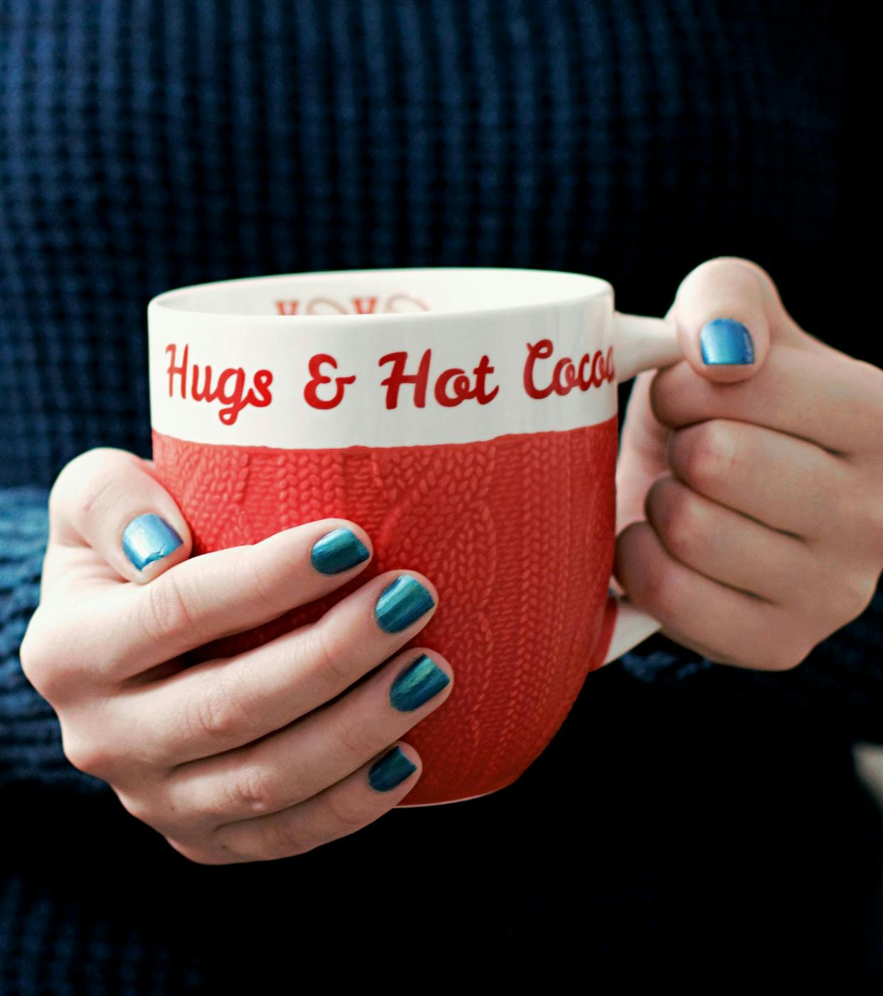  woman's hands with blue nail polish holding a red and white coffee mug with Hugs & Hot Cocoa written on it