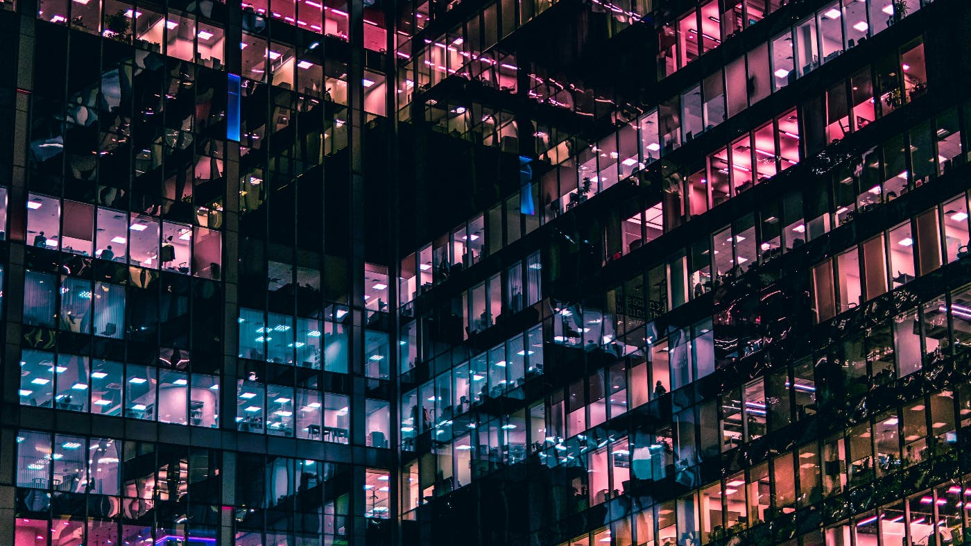 An architectural photo of a building at night with blue and purple lights and many people working inside