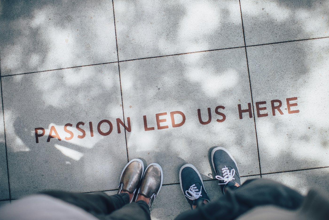 A sidewalk with the words Passion Led Us Here inlaid and two people's shoes