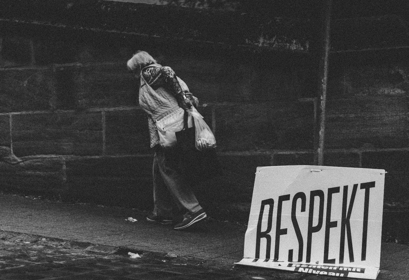 An older woman carrying groceries passes a sign falling off a pole reading RESPEKT