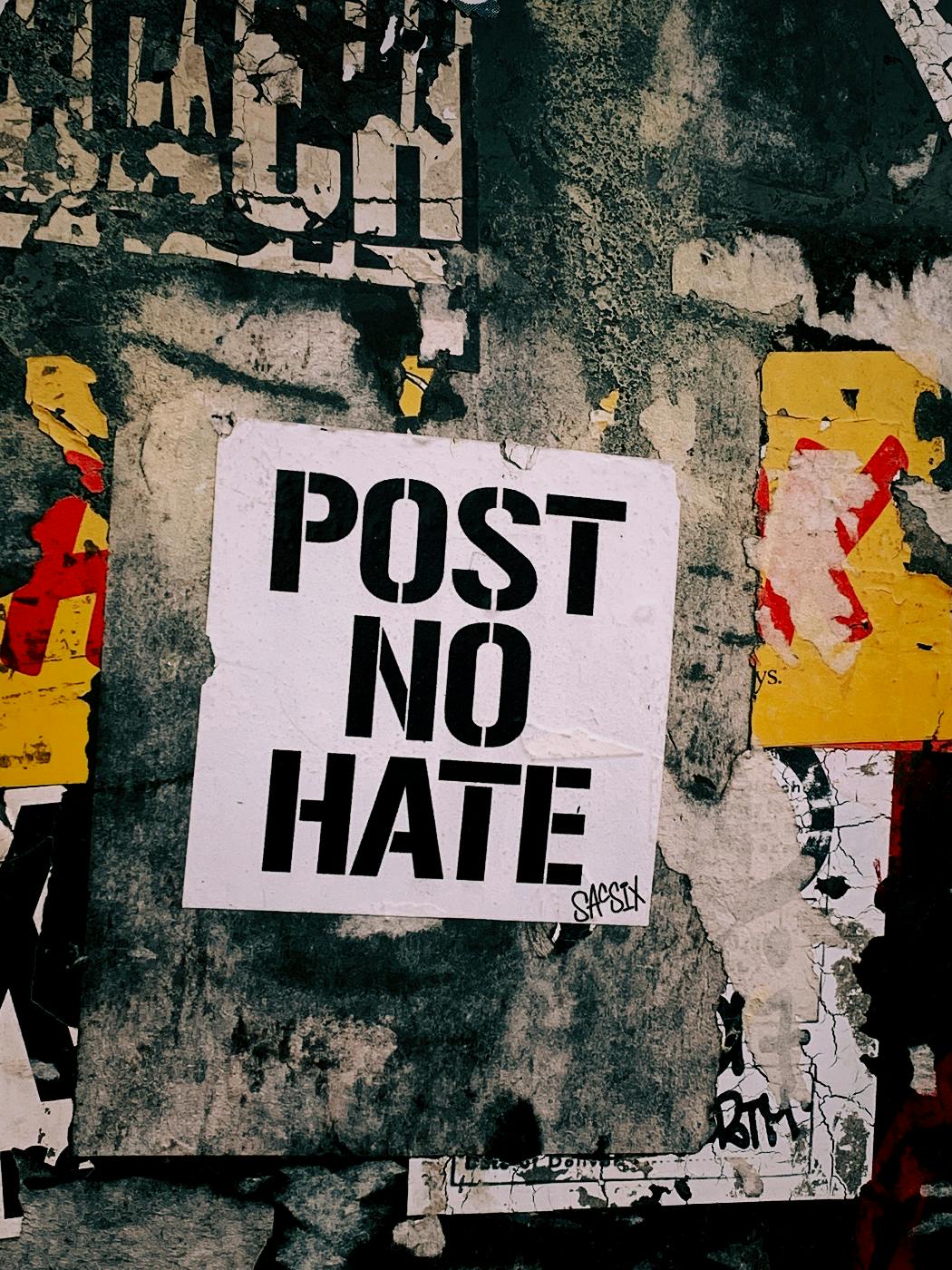 A graffiti covered wall with a bill in the middle reading POST NO HATE