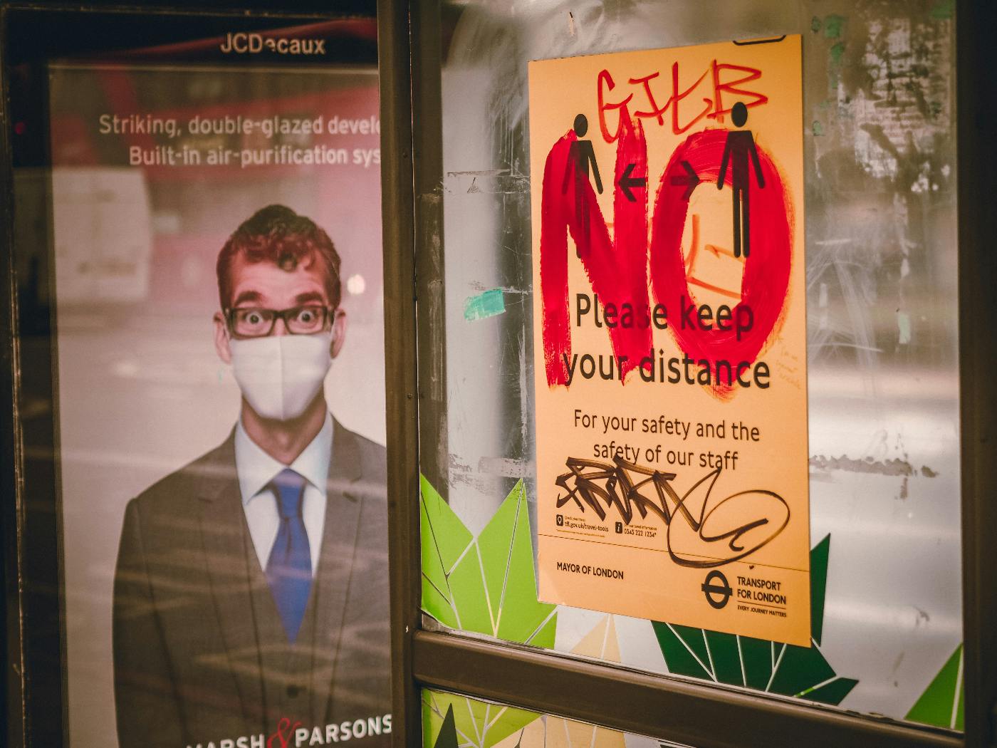A bus stop with poster ads: one shows a man wearing a mask, another asks fro sociladistancing which someone has written NO in red letters on