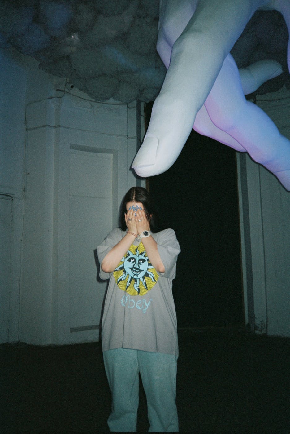 A woman with her hands covering her face and a giant hand coming out of the ceiling with the index finger pointing at her head.