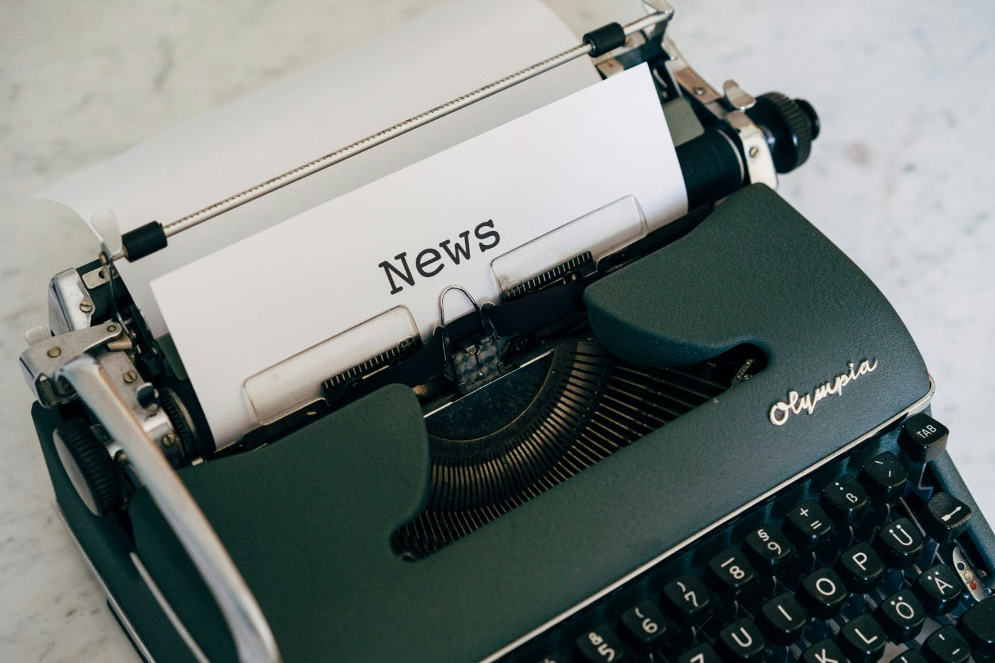A green Olympia typewriter with a sheet of paper in the roller with NEWS typed on it.