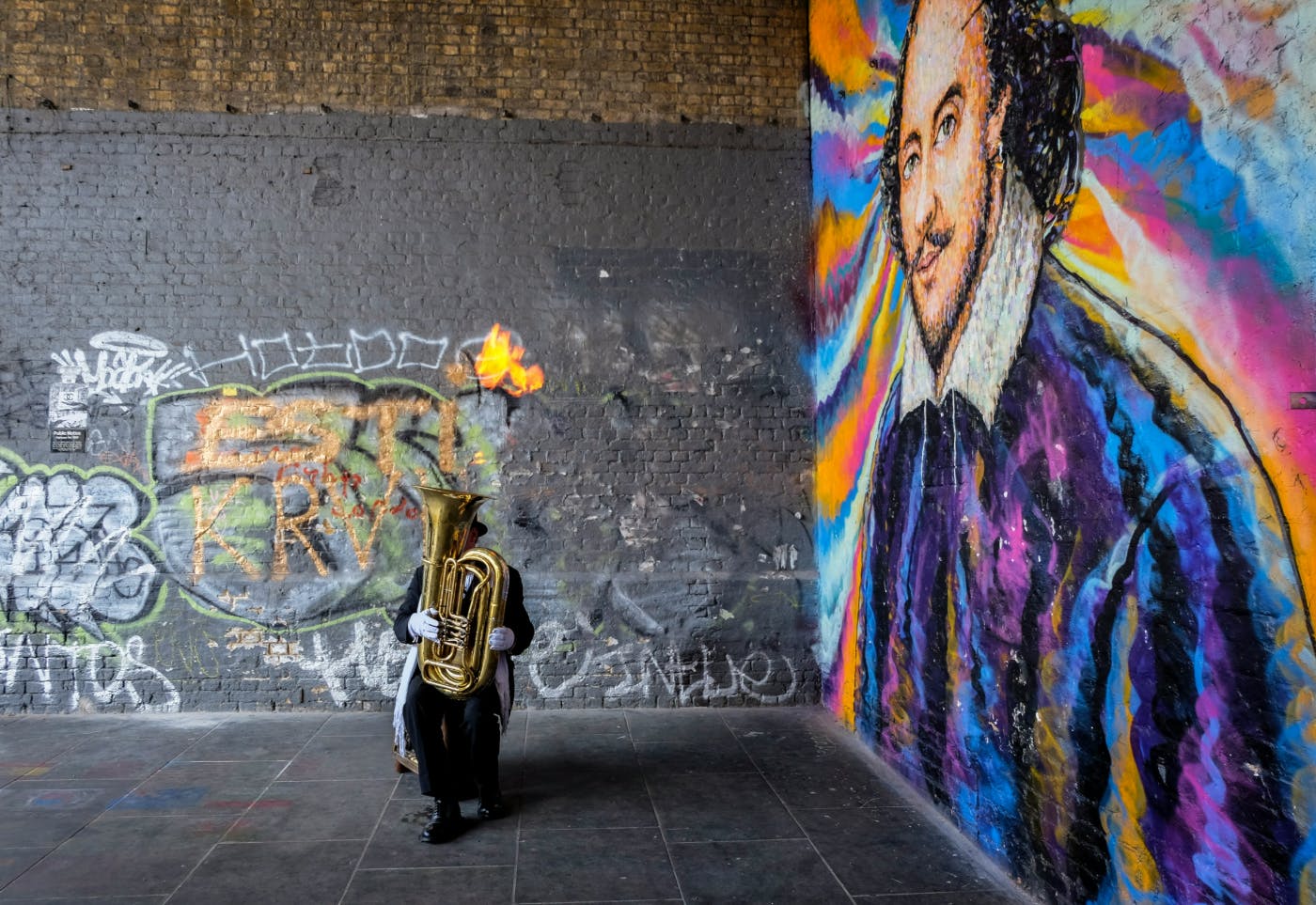 A man on the street in top hat and tails playing a tuba in front of a wall that has been graffitied with a portrait of Shakespeare in many colors