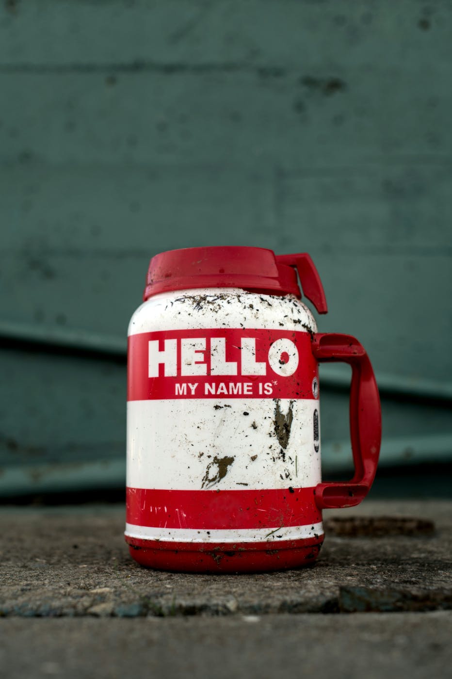A dirt cover red and white plastic travel coffee mug with Hello My Name Is on the side and then a blank space