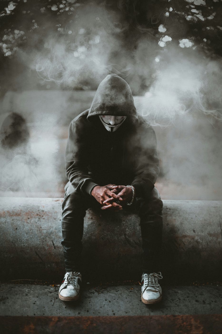 A man in a black hoodie and black pants wearing a V for Vendetta mask, sitting on the edge of a fountain