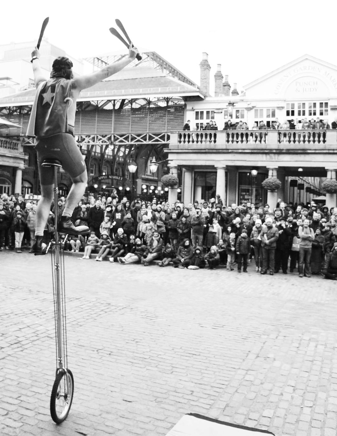 A street performer on a tall unicycle, holding knives in front of a big crowd