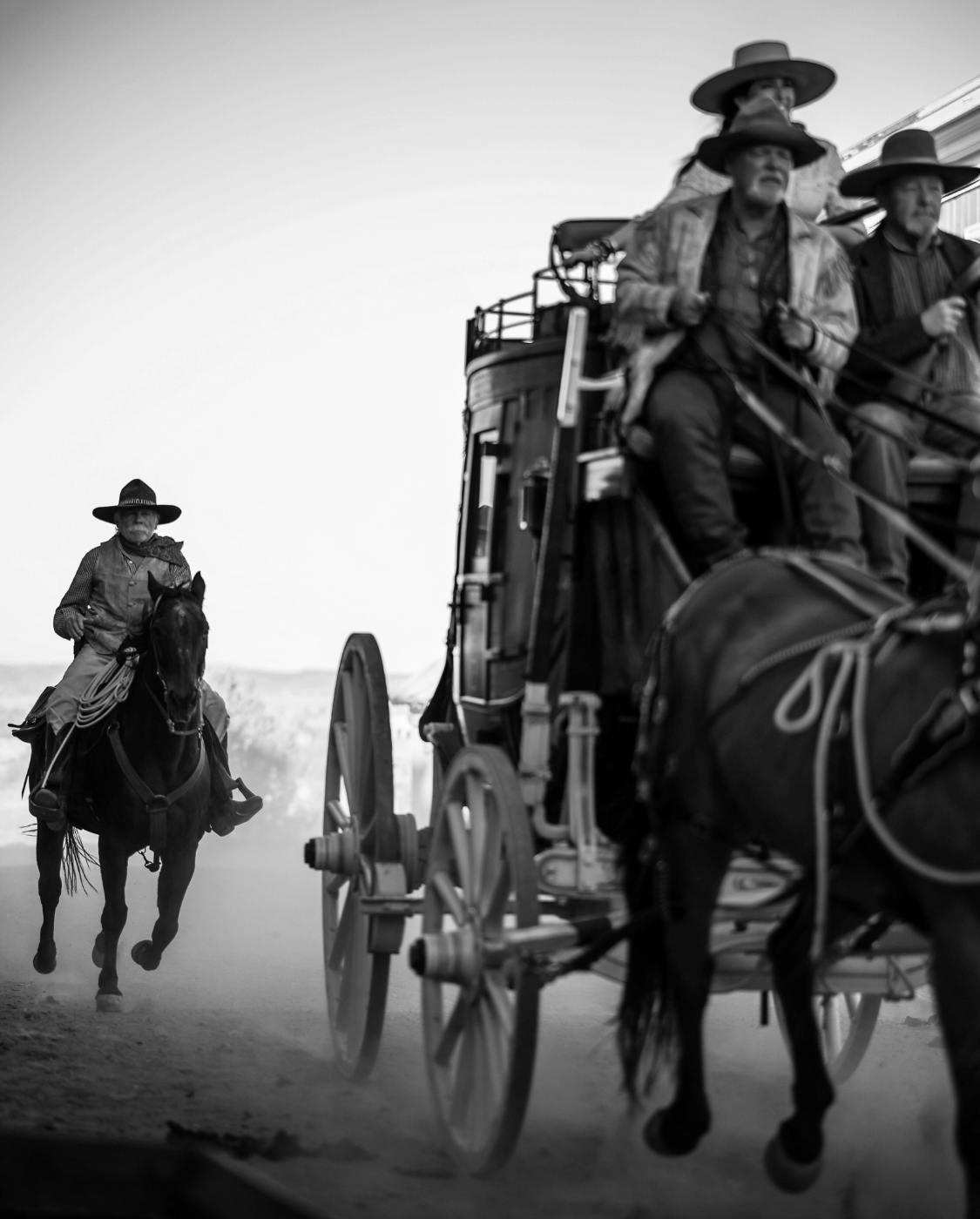 Three men on a stage coach with a rider following them