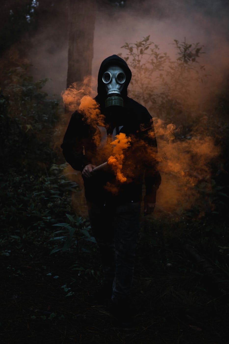 A man all in black wearing a a gas mask carrying and orange  smoke flare