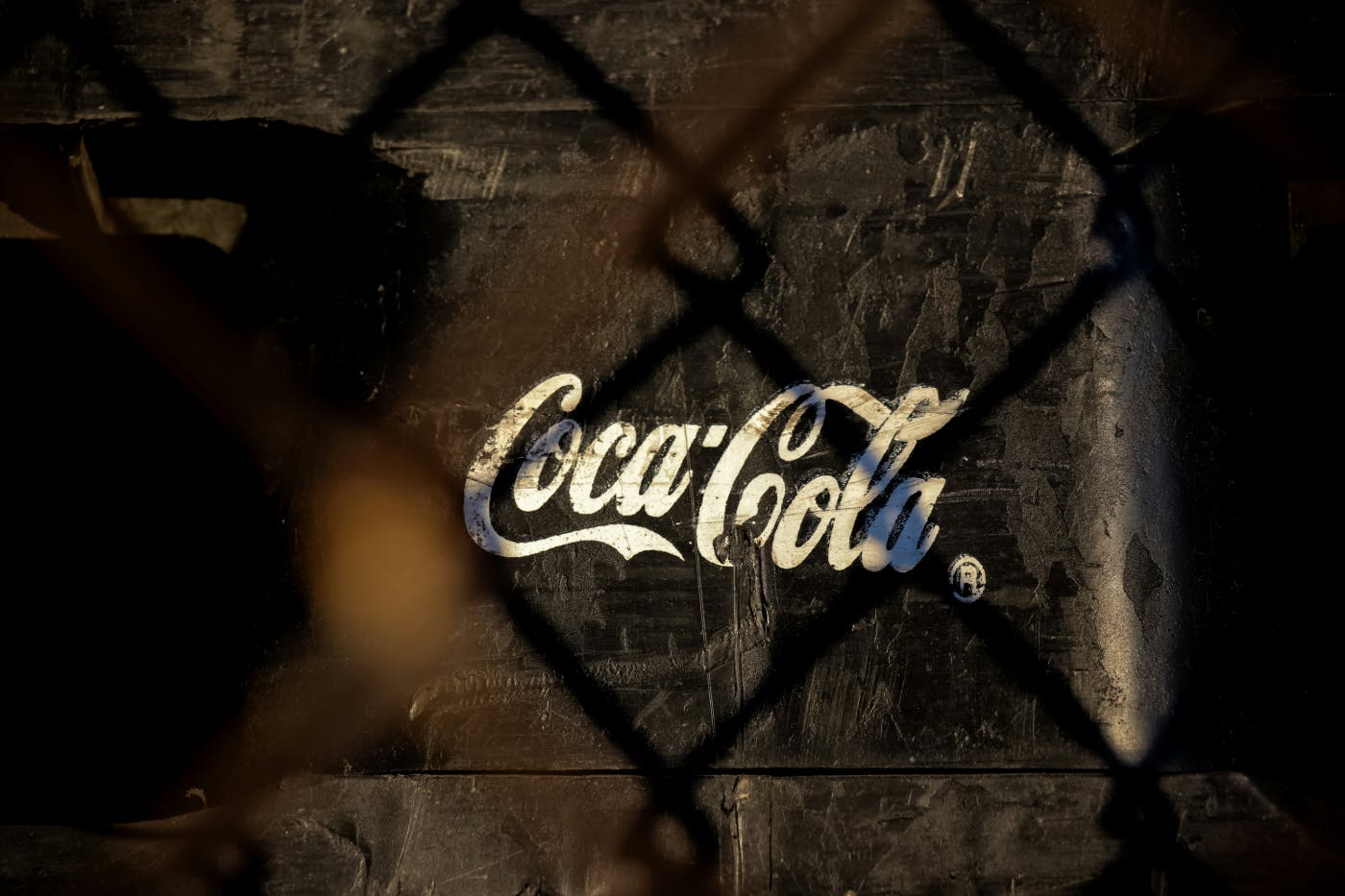 The Coca-Cola logo painted on a wall as seen through a chain link fence