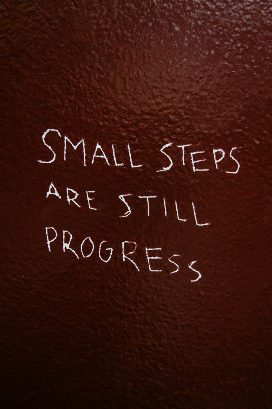 Letters scratched into a red painted wall reading, "Small Steps are still progress."