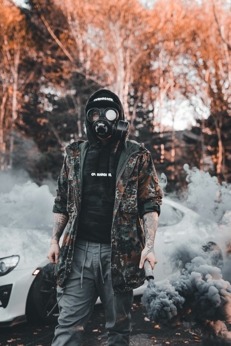 A man wearing a camo jacket and gas mask, carrying a smoke flare