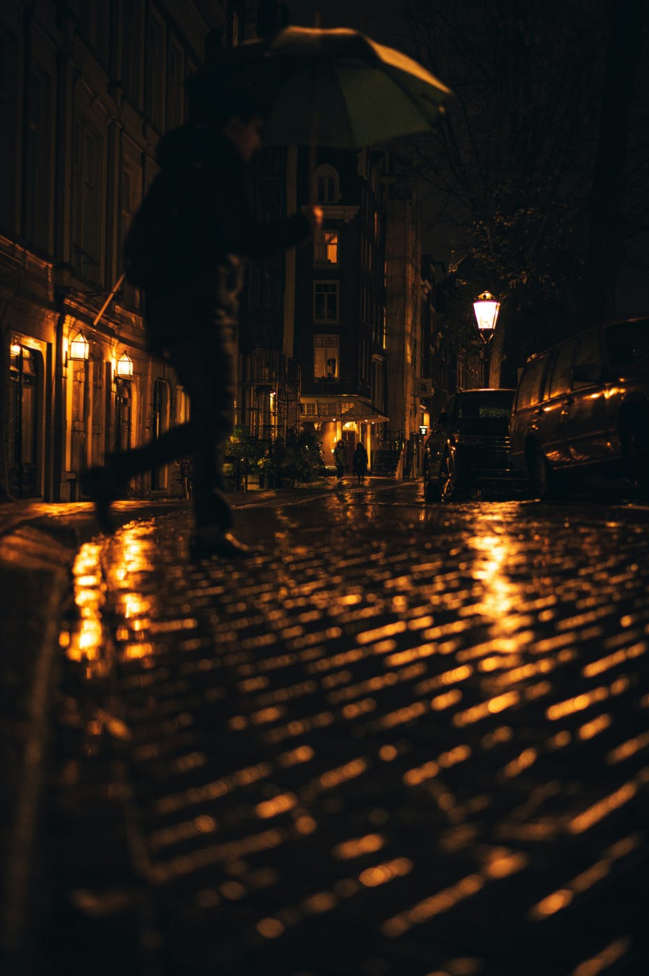 A barely visible man walks rainy cobblestone streets lit by street lamps