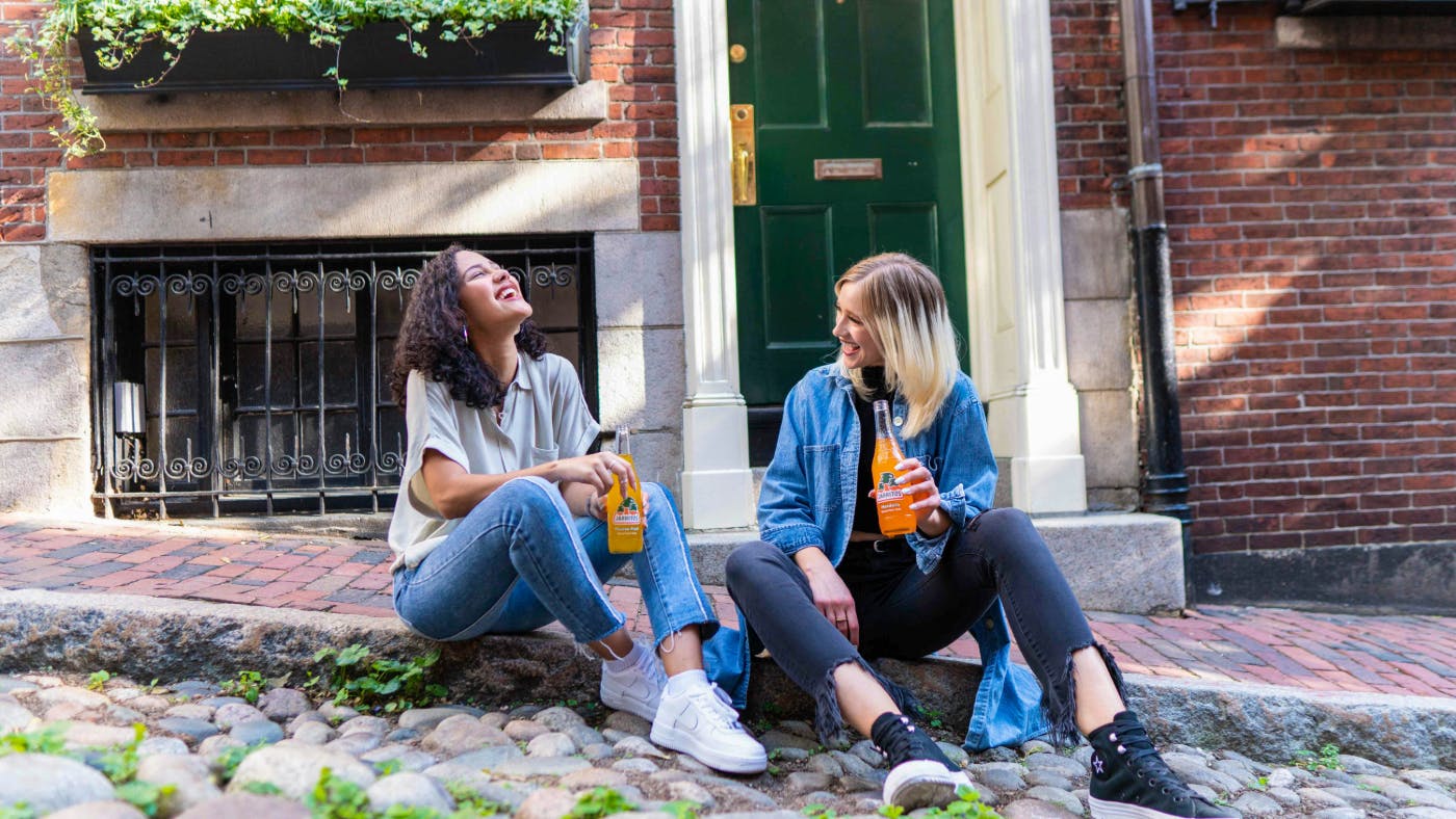 Two women sitting on a curb, drinking orange soda and laughing