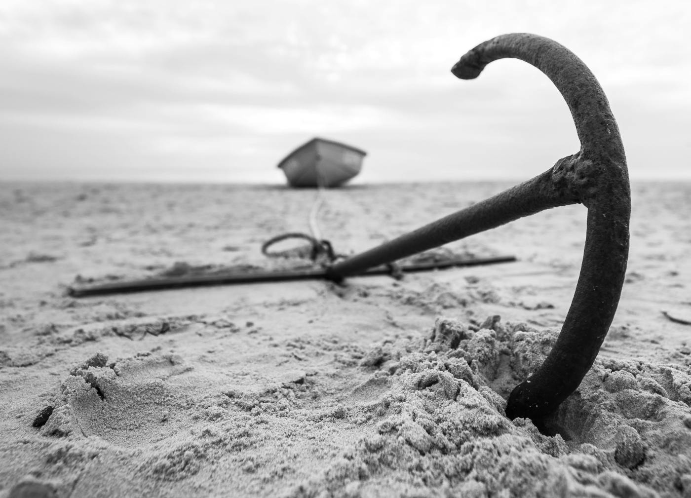 Gray scale, an anchor on the sand with a rope leading to a boat also on the sand