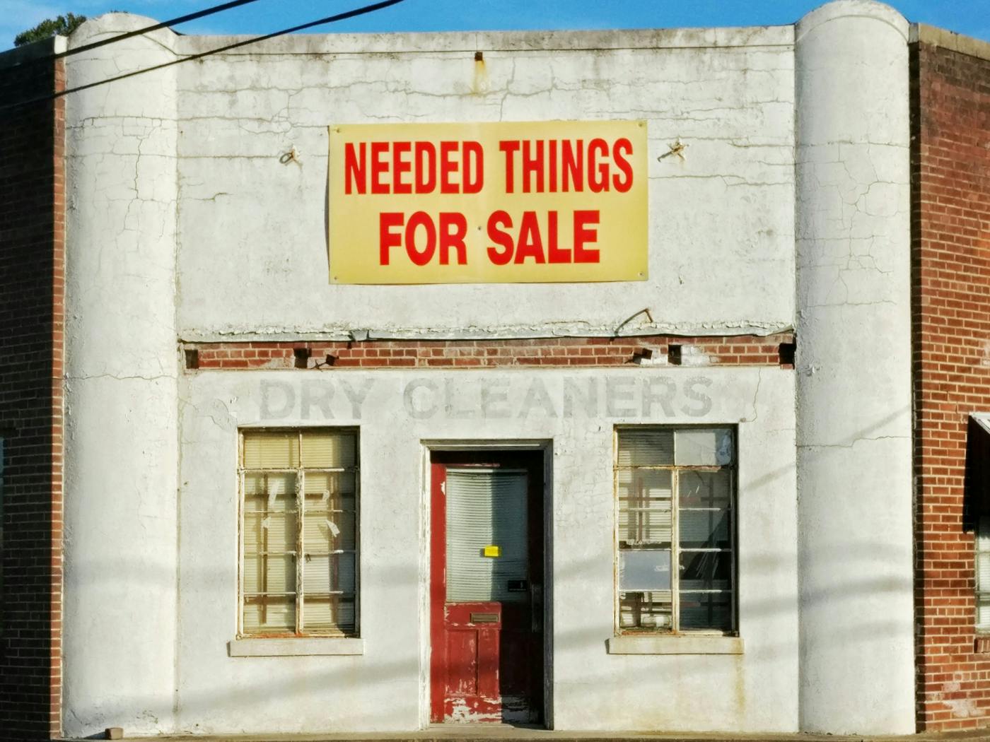 An old white building with a yellow and red sign reading Needed Things for Sale above the faded paint reading Dry Cleaners