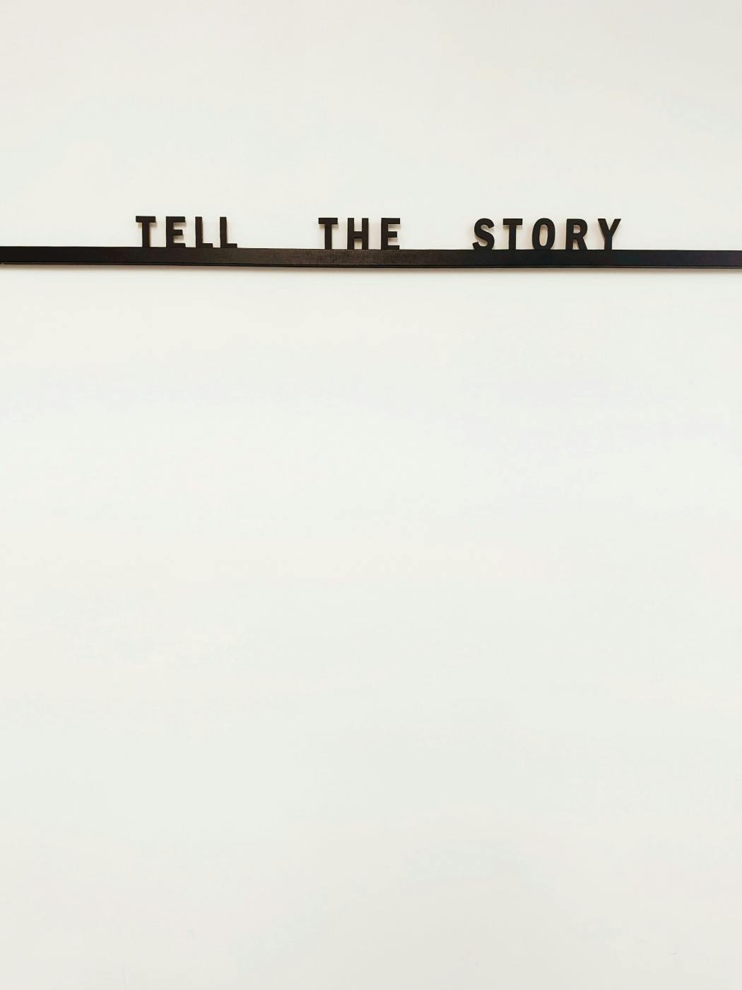 A blank wall with a thin self and the words Tell The Story on the shelf