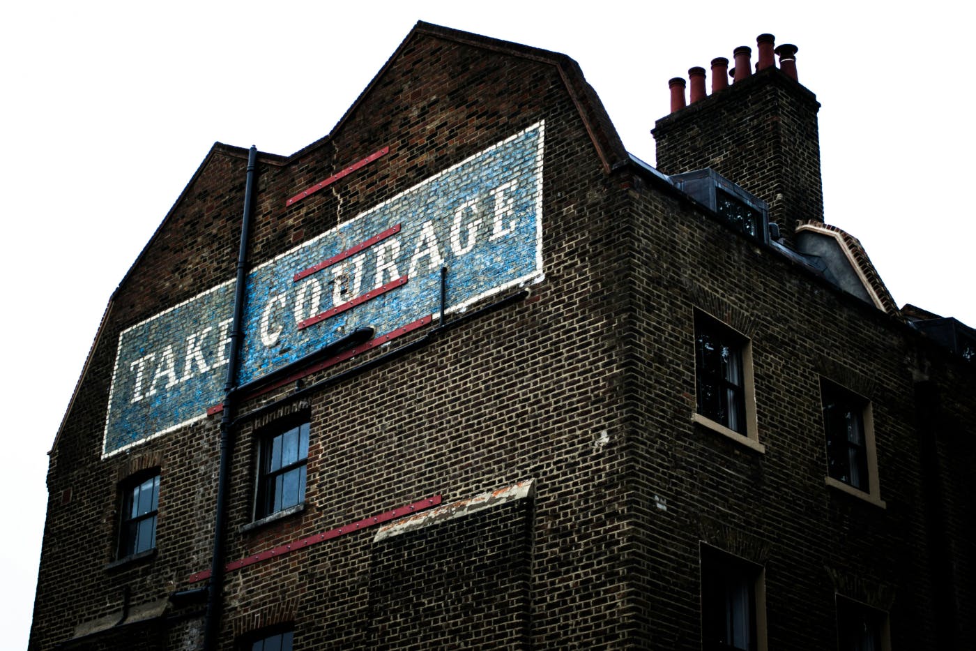 An old brick building with a blue and white sign painted on the side reading: Take Courage