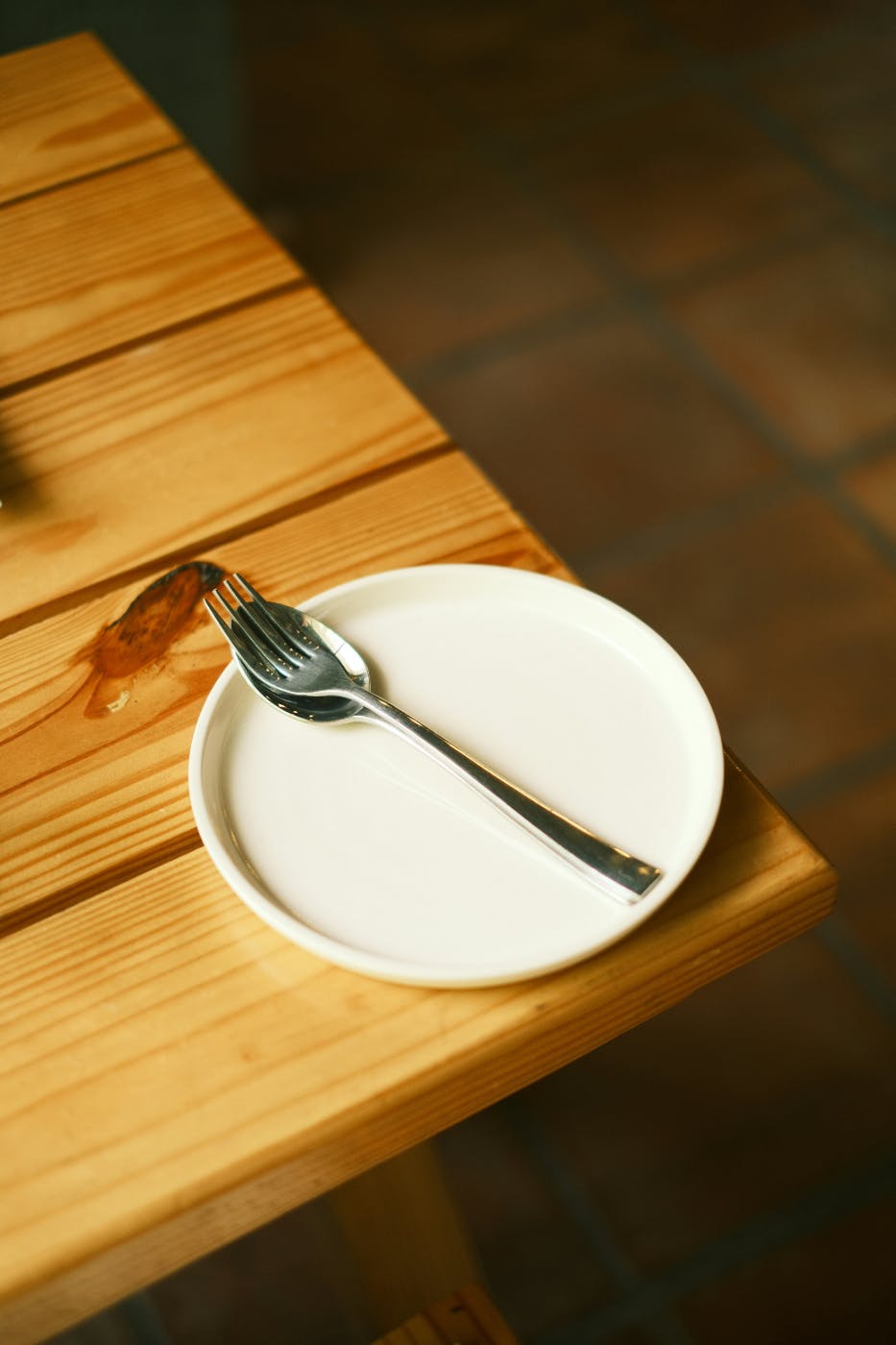 A white plate at the corner of a table with a fork and spoon on it. The fork on top of the spoon.