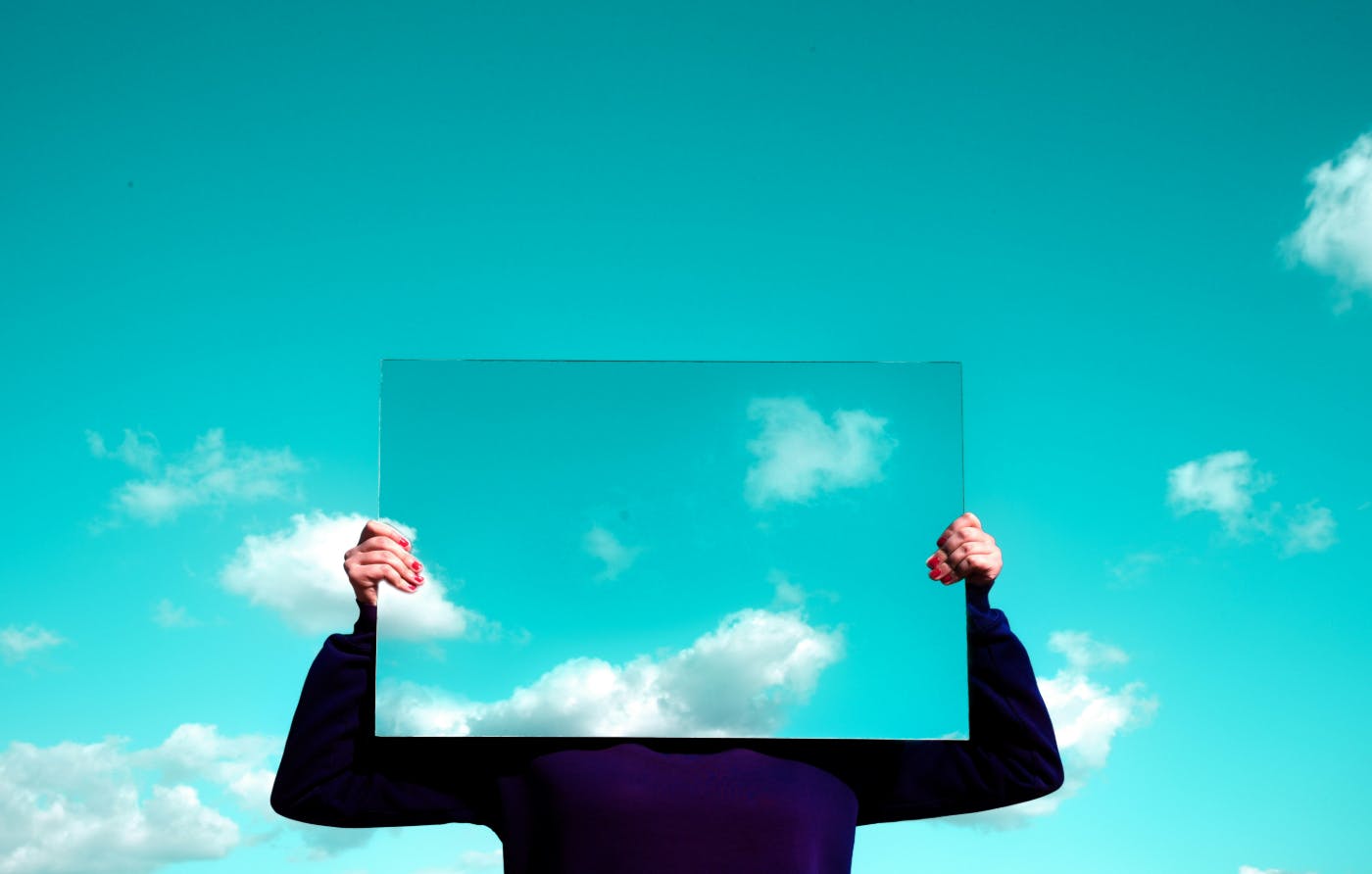 A person holding a mirror In front of their face that is reflecting blue sky and clouds