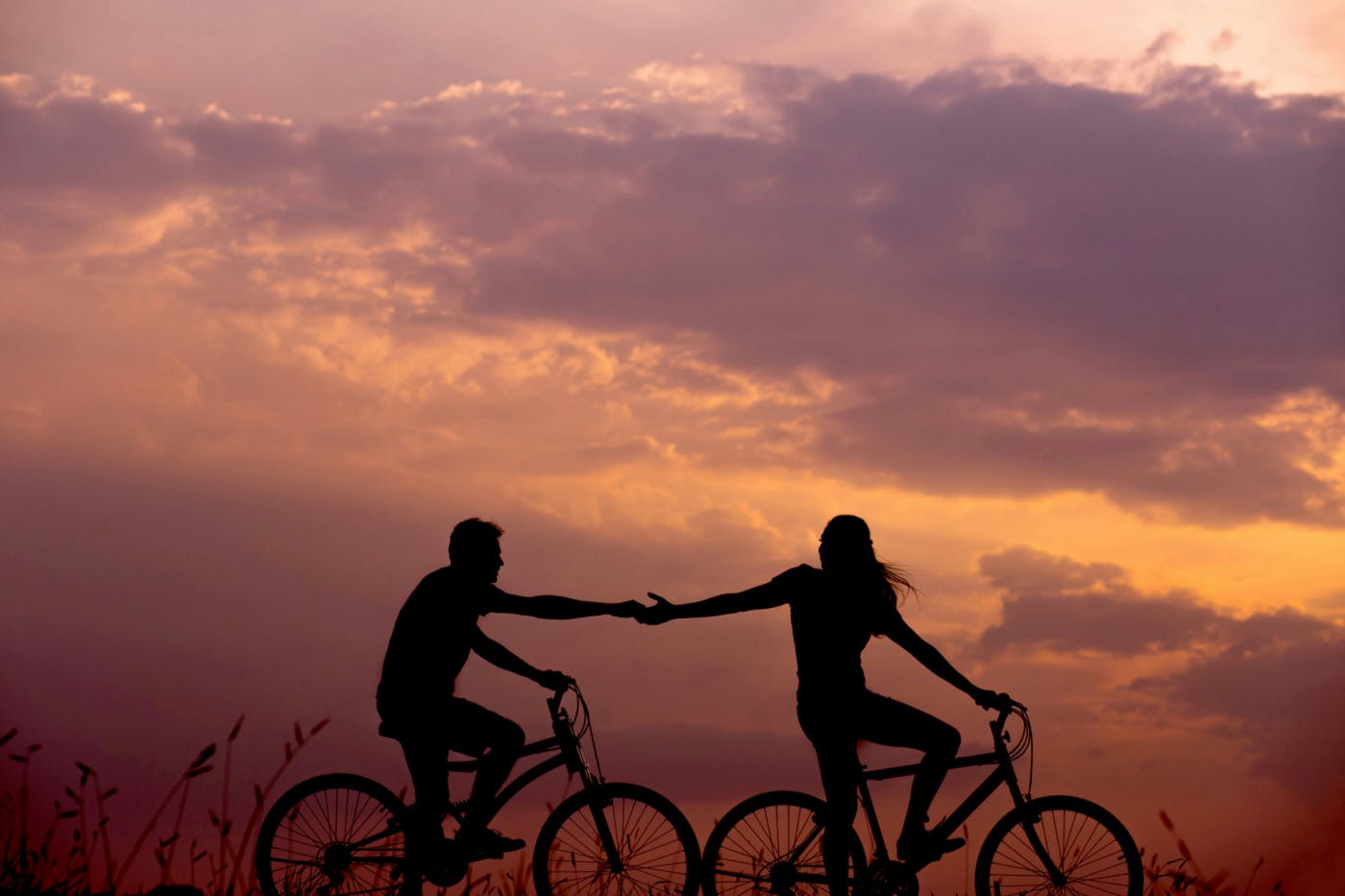 A woman on a bike reaching behind her for the hand of a man also on a bike against a sunset sky