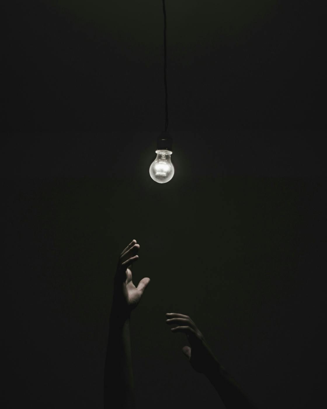 Two hands reaching for a lit bulb in darkness