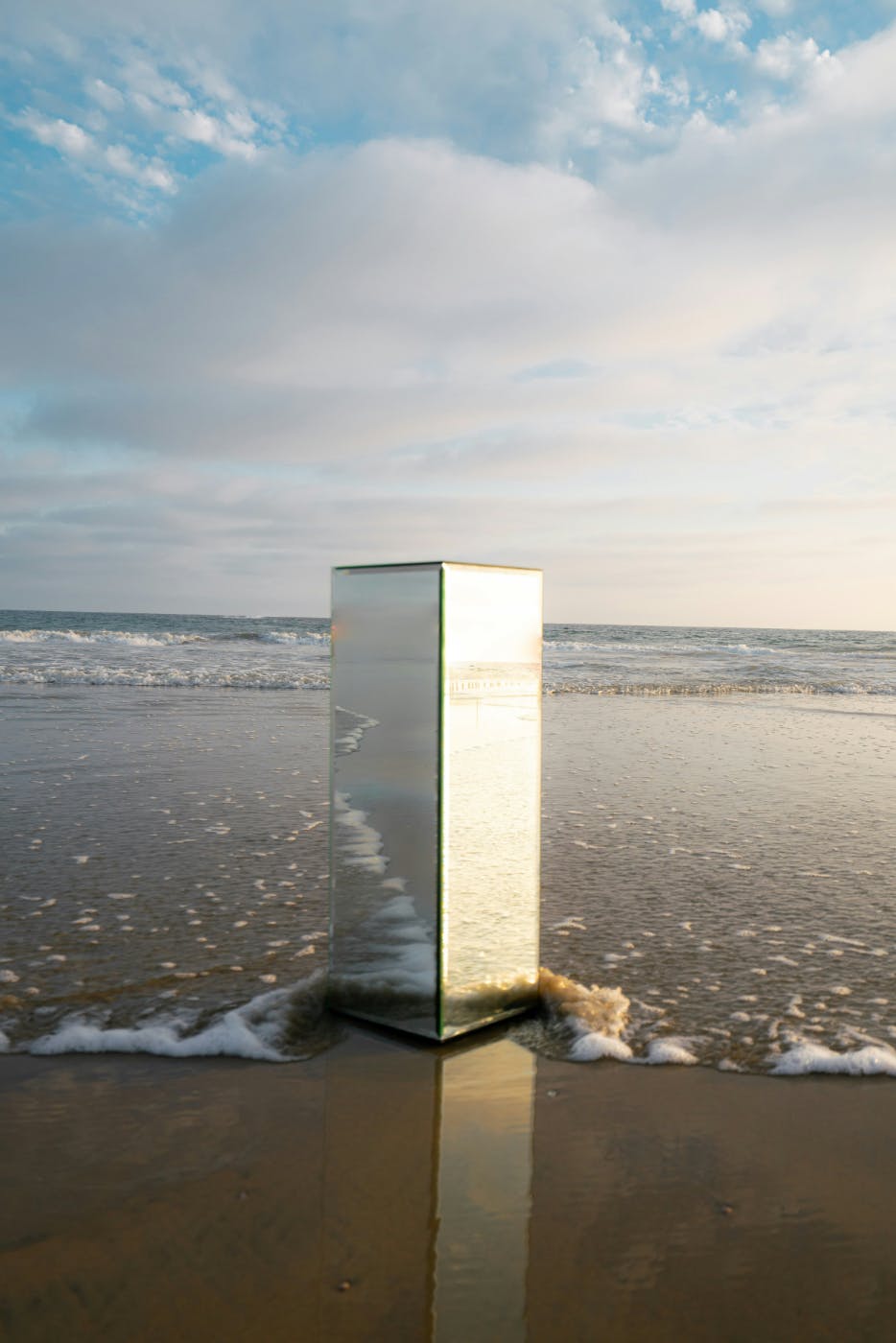 A square mirrored pillar sitting on the sand at surf's edge