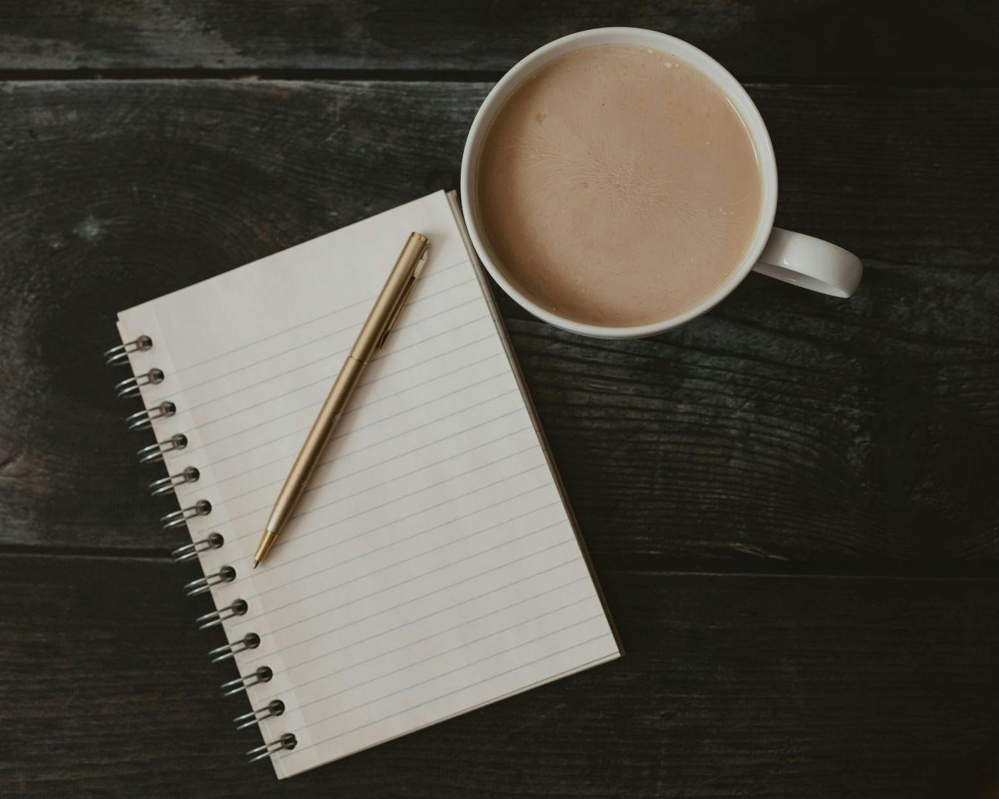 A blank white pad, a gold pen and a cup of coffee on a wood table