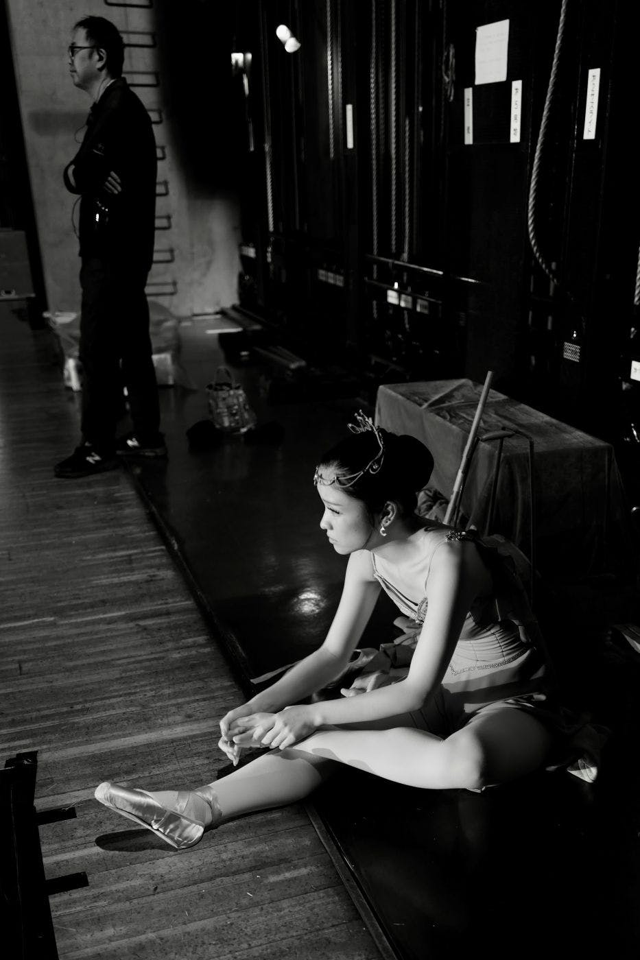 In the wings of a theater. A crew member watches the stage while ballerina adjusts her shoe