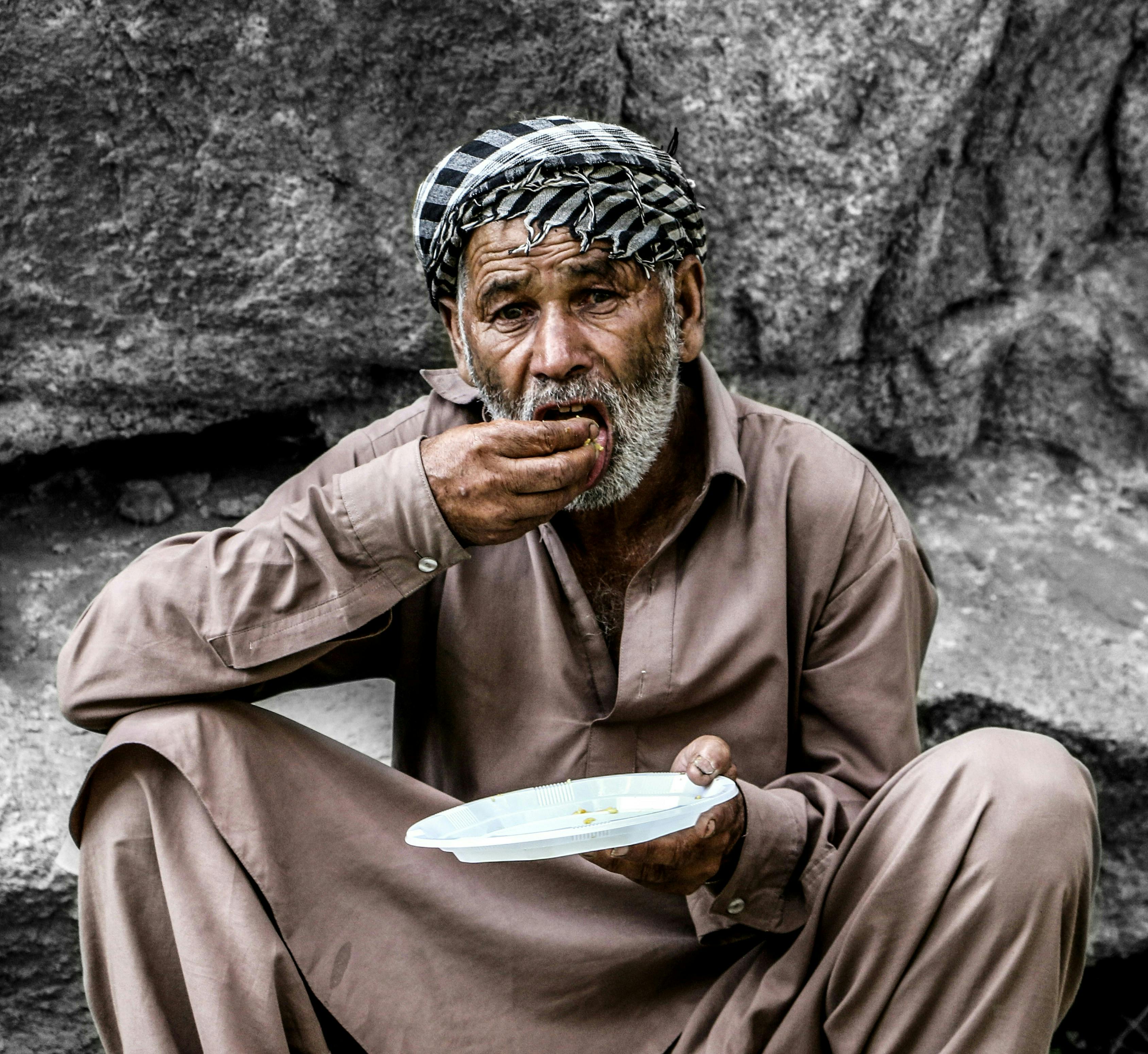 A man in a brown thobe sitting on the ground, eating from a plate with his hands