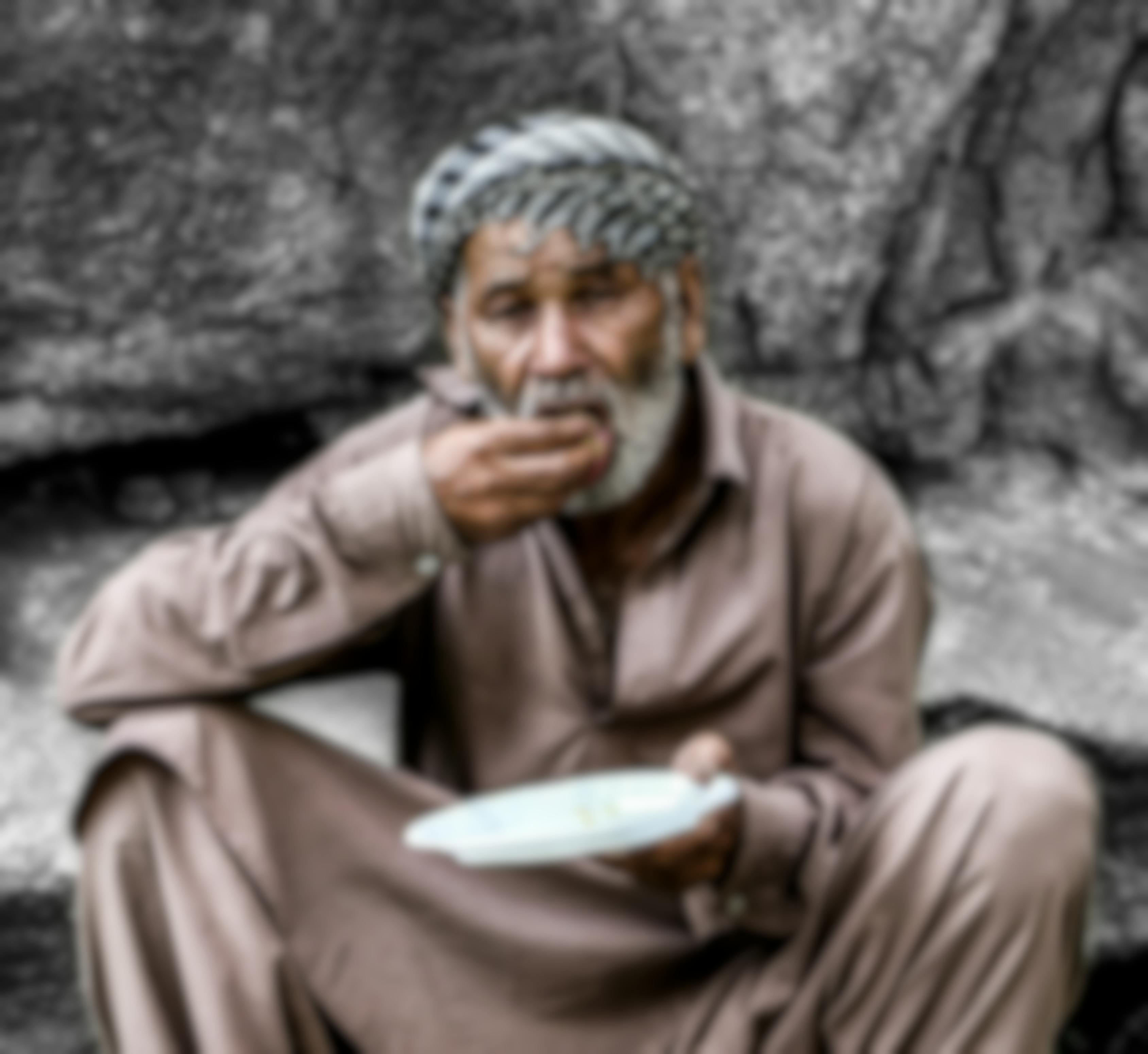 A man in a brown thobe sitting on the ground, eating from a plate with his hands