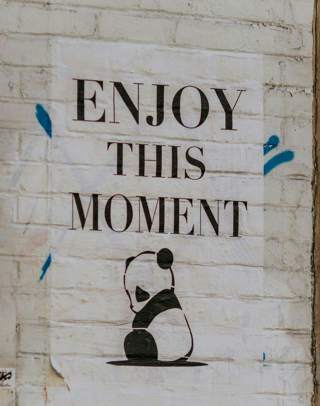 A poster on a white brick wall of a drawing of a panda with the words Enjoy This Moment above him
