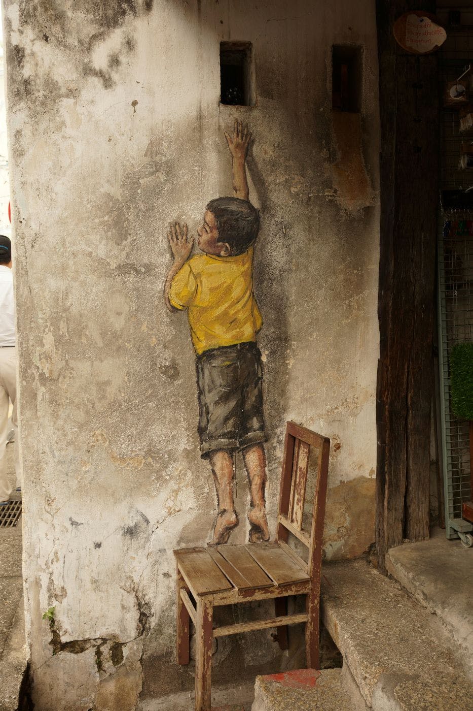 A wooded chair against a wall under the painting of a boy that looks like he's standing on the chaor