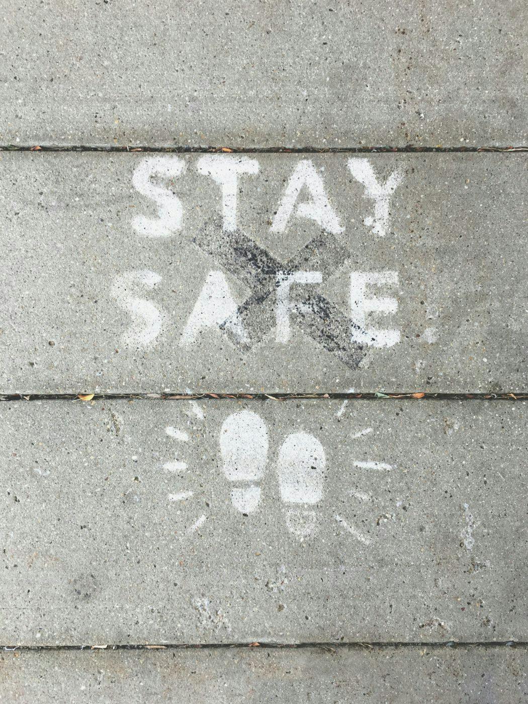 White letters stenciled on a sidewalk reading Stay Safe with two shoe prints