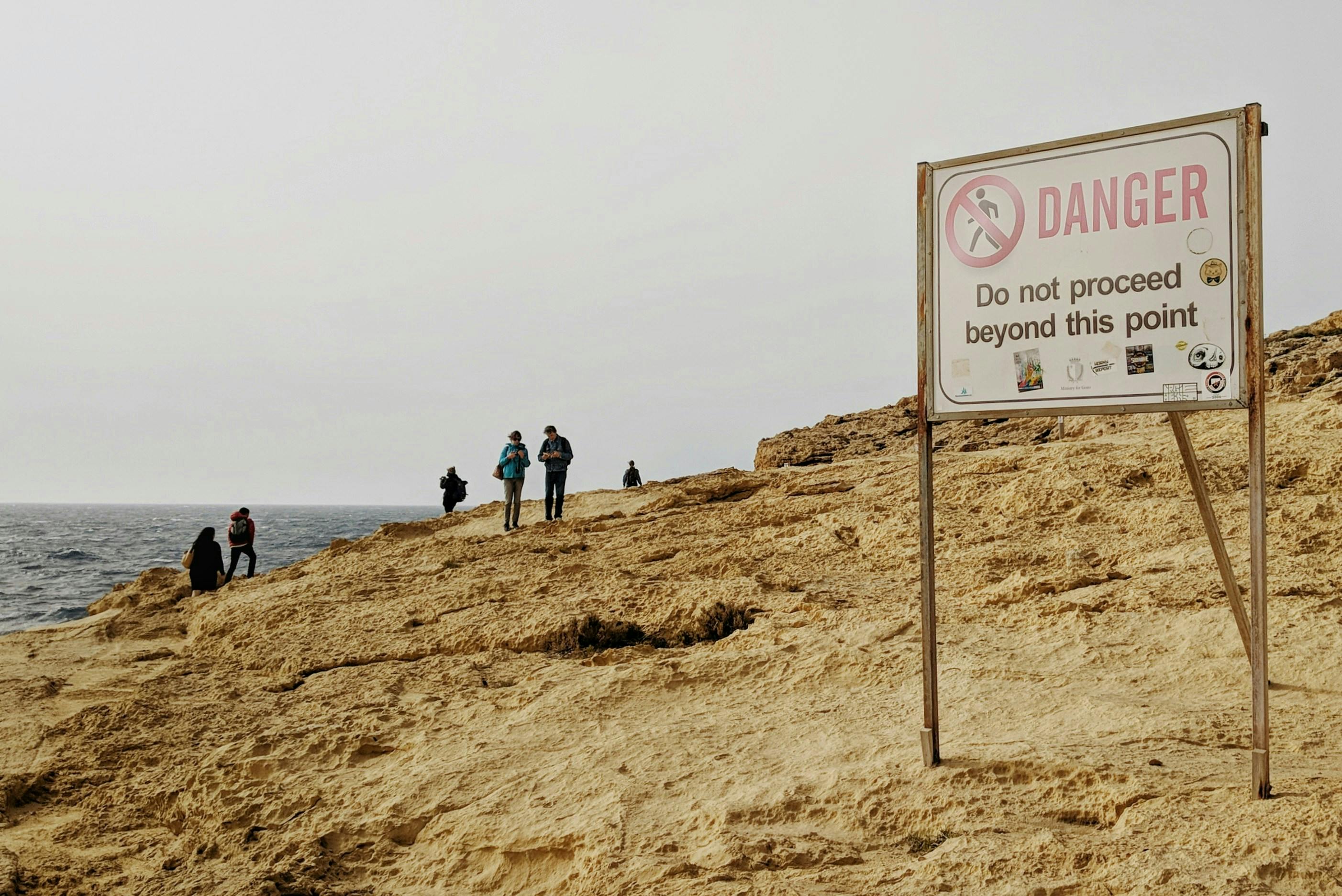 A sign on a beach reading Danger: Do Not Proceed Beyond This Point with many people beyond the point