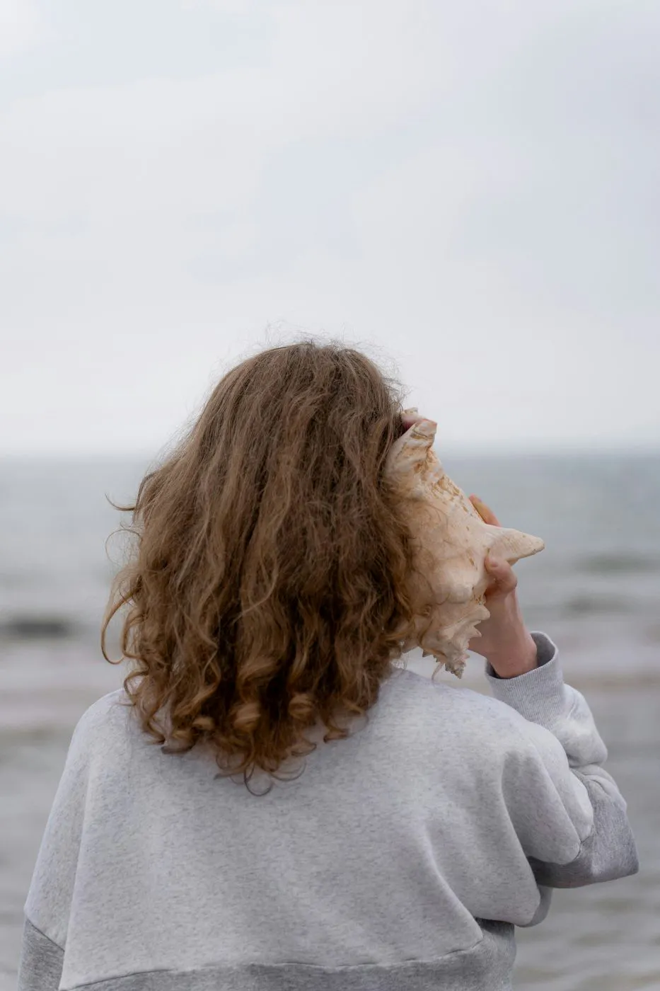 A woman by the ocean holding a conch shell to her ear