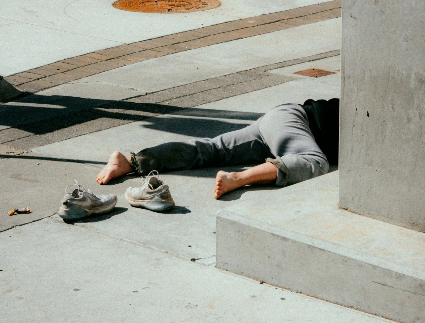 A man with shoes off, wearing sweatpants laying on the a sidewalk only half his body visible