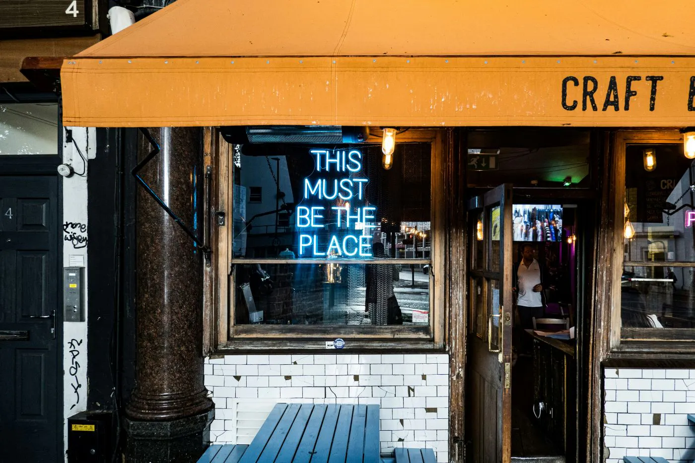 A small eatery with an orange canopy and a blue neon sign in a window reading This Must be The Place