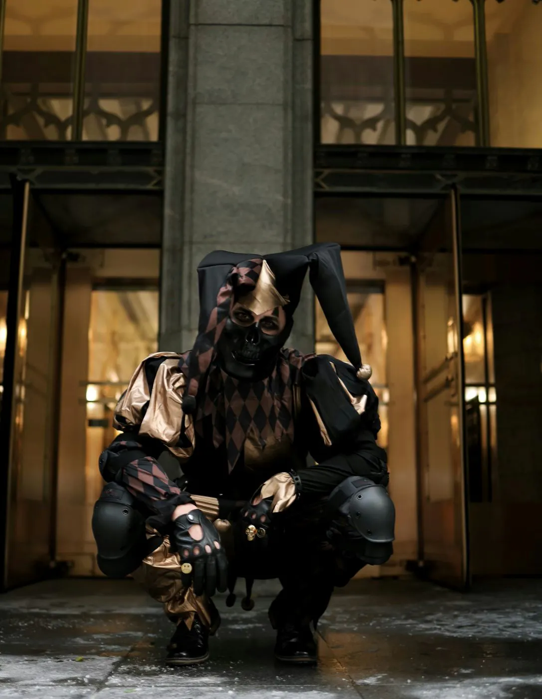 A person in black and gold motley crouching in front of a building