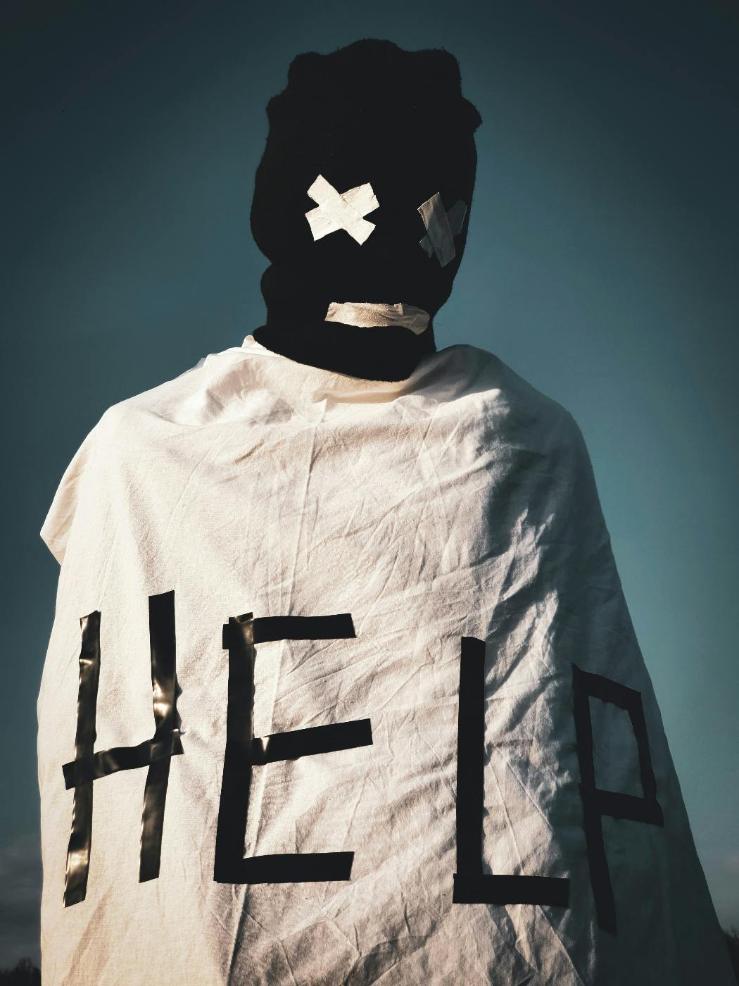 A person wearing a black ski mask and a sheet with the eye  and mouth holes taped shut and Help written on the sheet
