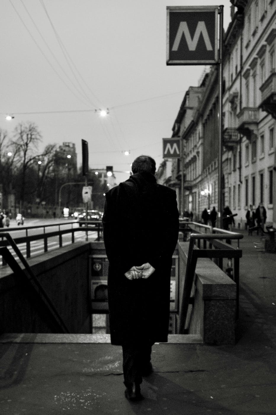 A black and white image of a man with hands behind his back entering a metro station