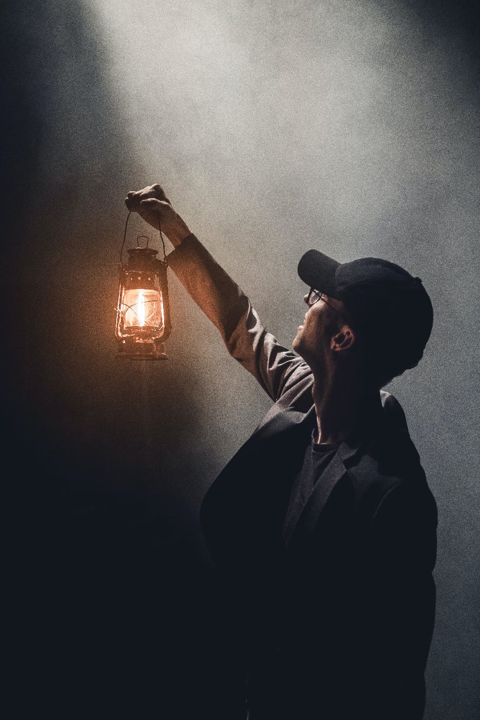 A man with a baseball cap in a beam of dusty light holding up a lantern